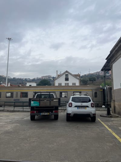 The image depicts an outdoor scene in Peso da Régua, Portugal, under an overcast sky. In the foreground, a concrete paved area serves as a parking lot. Two vehicles are parked, facing away from the viewer. On the left is a dark-colored pickup truck with a bed frame, displaying Portuguese license plate "36-ZP-78" and a teal sign for "Infraestruturas de Portugal" on its tailgate. To its right, a white Dacia Duster SUV, equipped with roof rails, shows Portuguese license plate "89-ZO-00". A yellow line delineating a parking space is visible on the right side of the concrete ground.

Immediately behind the parked vehicles, a stationary, light-colored passenger train with multiple carriages occupies a set of tracks. Visible text on the train includes "9101 19.980.313-3". Beyond the train, a white building with a dark-framed window and a gabled roof stands out. To the far right, a section of another white building with an overhang and patterned tiles is visible. A tall, grey street lamp post is positioned on the far left of the frame. The background consists of rolling hills extensively covered with residential and other structures, indicating an urbanized landscape. No individuals are present in the scene.