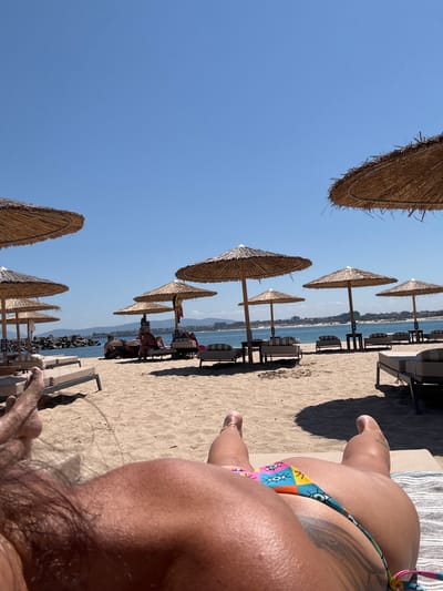 The image depicts a sunny day at a beach in Primorsko, Bulgaria. The scene is oriented vertically. In the foreground, the legs and torso of a person are visible, positioned on a sun lounger. The person is wearing a colorful bikini. The beach is sandy, with rows of straw umbrellas and sun loungers stretching towards the sea. The sea itself is visible in the background, with a coastline featuring buildings and some rocky structures. The sky is clear and blue, indicating fair weather. The time of day appears to be midday, given the bright sunlight and shadows. The overall atmosphere is one of relaxation and leisure, characteristic of a beach vacation.