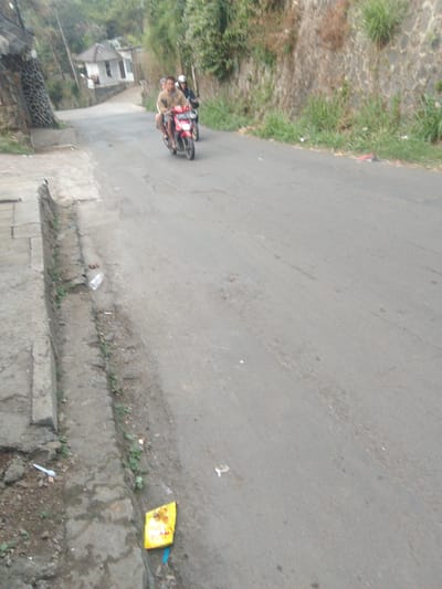 In Cisarua, Indonesia, two individuals are riding a red scooter on an asphalt road. The person in front is wearing a tan shirt, and the person behind them is wearing a black shirt and helmet. The scooter appears to be moving away from the viewer, up a slight incline.
The scene is a roadside on a hilly terrain. To the left of the road, there is a concrete ditch or drainage channel with some overgrowth of grass and weeds. Further to the left, a stone wall or structure is visible, along with some vegetation. Beyond the road on the right, a steep, rocky embankment covered in grass and some trees rises. In the background, a small building with a light-colored roof and walls can be seen nestled among trees, suggesting a residential area on the hillside.
The overall lighting suggests it is daytime, possibly late morning or early afternoon, with no strong shadows indicating harsh sunlight. The weather appears to be clear and mild, with abundant greenery suggesting a tropical climate.
Notable details include some litter on the side of the road, specifically a yellow package with some red markings near the foreground and a clear plastic bottle in the ditch. The road surface shows signs of wear. The image quality is somewhat blurry, possibly due to motion or low resolution. No discernible text is visible on any of the objects or surroundings. The atmosphere appears to be ordinary, depicting a typical day in a rural or semi-rural Indonesian setting.