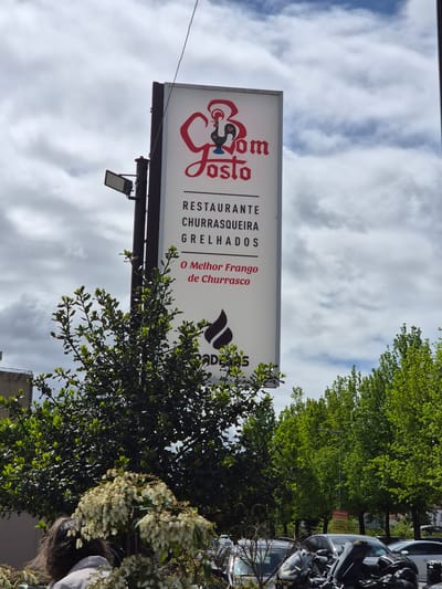 The image features a tall, rectangular sign for "Bom Gosto," a restaurant in Barcelos, Portugal. The sign, colored in white and red, lists "Restaurante," "Churrasqueira," and "Grelhados," along with the phrase "O Melhor Frango de Churrasco." The sign also shows the logo of "ADERS."

The sign is erected on a pole, positioned against a sky filled with scattered clouds. Lush green trees surround the sign, partly obscuring it. The background also contains parked cars and glimpses of buildings. A woman is visible at the bottom of the image, standing near flowers and a vehicle.