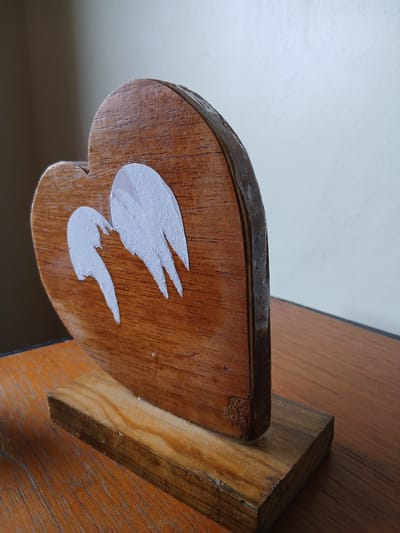 A close-up, eye-level, medium shot captures a wooden heart-shaped ornament with two white angel wings adhered to its surface. The ornament stands upright on a wooden base, which rests on a wooden table. The background consists of a wall, with the top portion appearing lighter than the bottom. The ornament is positioned slightly to the left of the frame's center. No people are present, and no specific actions or interactions are depicted. The scene does not provide any discernible information about its location in El Valle del Espíritu Santo, Venezuela.