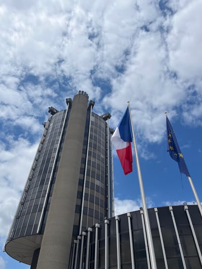 The image shows a low-angle view of a modern skyscraper in Créteil, France, under a partly cloudy sky. The building features a dark, reflective glass facade and a prominent cylindrical concrete tower with vertical ribbed detailing. Two flags are visible on flagpoles in the foreground: the French tricolor and the European Union flag. The French flag, with its blue, white, and red vertical stripes, is positioned to the left, while the blue EU flag with its circle of yellow stars is to the right. The flags are illuminated by natural daylight. The architecture suggests an institutional or governmental building. The environment is urban, with the focus on the building and the flags against the backdrop of the sky.