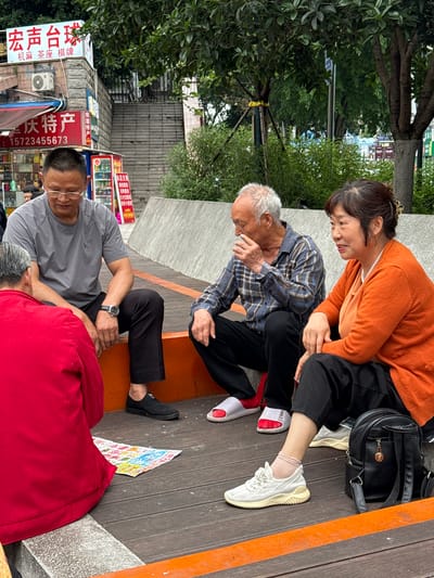 Here is a description of the image:
**Location:** Nan'an District, China. This is inferred from the visible Chinese signage.
**Scene Description:** The image shows a street scene where several people are interacting.
**People:**
* Two older people, a man and a woman, are seated on a raised wooden platform. The woman is wearing an orange sweater and black pants. The man is wearing a plaid shirt and black pants. The woman appears to be attending to the man, who is holding his neck or head.
* Two additional men are on the ground. One is wearing a grey shirt and black pants, and appears to be looking at or tending to the other, who is lying on his back and partially covered by a red blanket.
**Objects:**
* A black bag is next to the woman.
* A flyer or printed material is on the wooden platform.
* Various storefronts and signage are visible in the background, suggesting a commercial area.
* An air conditioning unit is attached to one of the buildings.
**Actions and Interactions:**
* The woman appears to be attending to the man seated next to her.
* The man in the grey shirt appears to be tending to the man lying on the ground.
**Environment:**
* The scene is outdoors, likely on a public street.
* The presence of trees and buildings indicates an urban or semi-urban setting.
* The wooden platform suggests a designated seating area or public space.