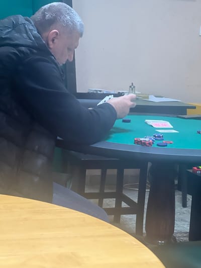 An indoor scene within the city of Smederevska Palanka Municipality, Serbia, depicts a male individual engaged in a card game. The subject, a male with short, graying hair, is observed in profile, facing right, wearing a dark, possibly quilted, jacket and dark trousers. He is seated at a dark-rimmed poker table featuring a green felt playing surface. His left hand holds a fanned hand of multiple playing cards, while his right hand is positioned near the cards. On the green felt, several poker chips are visible, including stacks of red and black chips, and individual blue and purple chips. Additionally, a stack of face-up playing cards and some face-down cards are present on the felt. In the background, behind the poker table, a dark elevated surface holds a clear glass shaker and several papers. The surrounding environment includes a light beige wall and a section of a dark teal wall to the left. The floor is covered with light-colored tiles. A light wooden table is partially visible in the foreground, obscuring the lower portion of the subject and the poker table. A white electrical cable is discernible on the tiled floor beneath the tables.