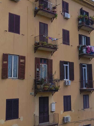 This is an exterior, eye-level, medium shot of a multi-story building with a light yellow facade. The building has numerous windows and balconies. The windows are mostly shuttered with dark purple or brown wooden shutters, some of which are open. The balconies have black metal railings and are adorned with potted plants. On some balconies, laundry is hanging out to dry. Two air conditioning units are visible on the exterior wall. The building appears to be residential, and the style suggests it is located in a European city. The specific location is in Rome, Italy, based on context not available in the image itself. There are no people visible in the image. The lighting is neutral, indicating an overcast or diffused daylight.