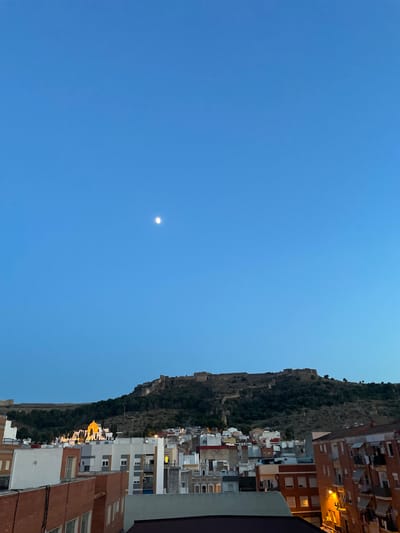 The image shows a panoramic view of the city of Sagunto, Spain, from a rooftop perspective. In the foreground, several residential buildings are visible, characterized by their varied architecture, colors, and balconies. Some windows are illuminated, suggesting it is dusk or evening. The middle ground is dominated by a densely populated urban area with more buildings, a prominent yellow dome of a church or religious building, and trees. In the background, a large, ancient castle complex is situated atop a hill, overlooking the city. The sky is a clear, deep blue, with a small, bright moon visible in the upper portion of the frame. The overall scene depicts a historical Spanish city at twilight.