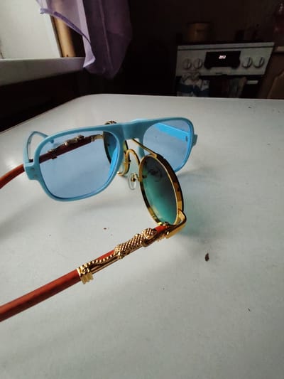 The image shows a pair of blue-rimmed sunglasses with blue lenses and gold accents on a white surface. One lens is in focus, showing a reflection of a gold, ornate temple arm. The other pair of sunglasses is slightly out of focus, with a blue frame and blue lenses. The background is blurred but suggests an indoor setting with a kitchen appliance visible. There is no indication of the city of Чухлэм, Russia.