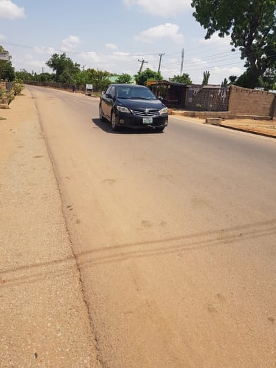 The image shows a black car parked on a paved road in Palade, Nigeria. The car is positioned on the left side of the frame, facing slightly towards the viewer. The road appears to be made of asphalt with some gravel on the shoulder.

In the background, there are buildings, trees, and utility poles with electrical wires. The sky is blue with some white clouds, suggesting it is daytime and the weather is fair. A fence can be seen along the side of the road, bordering some of the properties. One of the buildings has what appears to be a small shop or stall with metal bars on its windows.

There is a sign visible on the side of the road with some text, but it is too small to read clearly. The overall scene depicts a typical street in a populated area, likely a town or city, in Nigeria. The lighting suggests it is a bright, sunny day. No people are clearly visible in the image, though there might be a person in the distance near the buildings. The image is oriented vertically.
