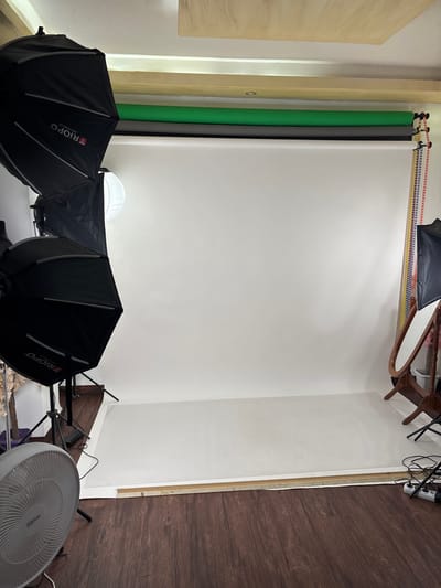 This image shows a photography studio setup in Amman, Jordan. The scene features a backdrop with several rolls of colored paper, including green, gray, and white, hanging from a rack. In the foreground, professional lighting equipment, specifically two large softbox umbrellas with the "TRIOPO" logo visible on one, are positioned on stands. A white, dome-shaped diffuser is also present. The floor is covered with dark wood laminate flooring, and a white oscillating fan is visible on the right side of the frame. The walls are painted white, and a wooden door frame is partially visible on the left. The lighting suggests an indoor setting, likely during the daytime given the overall illumination, although no windows are clearly visible. The arrangement of equipment indicates that a photo shoot is either in progress or has recently concluded, but no people are present in the image. The overall impression is one of a functional and organized workspace for photography.