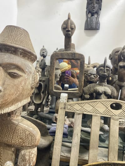 The image depicts an interior space, identified as being in Alaguntan, Nigeria, densely populated with traditional carved wooden artifacts. In the left foreground, a large, light-brown, weathered wooden sculpture of a human figure, possibly male, is prominently positioned. This figure wears a patterned, wide-brimmed hat, and features a detailed, carved left eye.
Centrally in the mid-ground, a rectangular, intricately carved wooden mirror reflects a person holding a purple-cased smartphone. The individual in the reflection possesses dark skin, wears a light-colored, possibly yellow, knitted hat with a ruffled brim, and a dark top. This reflection indicates the photographer.
Surrounding the central mirror and extending into the background are dozens of additional dark-toned wooden sculptures. These include various masks, busts, and full figures, many exhibiting distinct human or stylized features. Several darker wooden masks are mounted directly onto a plain white wall in the upper background. To the right side of the mid-ground, a section of a light-colored carved wooden chair, featuring geometric patterns on its backrest, is partially visible. The overall environment suggests a collection or retail display of traditional African art.