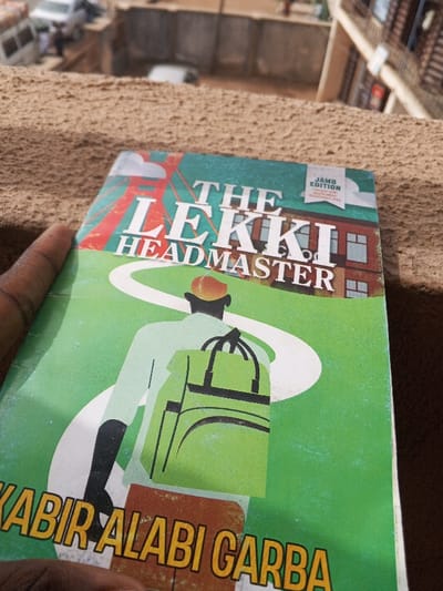 A dark-skinned hand holds a copy of the book "THE LEKKI HEADMASTER" by Kabir Alabi Garba, displaying its green, red, and white cover which features an illustration of a person with an orange cap and a green backpack. A "JAMB EDITION" label is visible on the top right of the book's cover. The hand and book are positioned above a textured, sandy-brown parapet, suggesting an elevated viewpoint within the city of Oko Erin, Nigeria. In the blurred background, an urban street scene is visible, featuring a white minibus and other vehicles on a road, with multi-story buildings and faint human figures in the distance. The scene is illuminated by natural daylight.