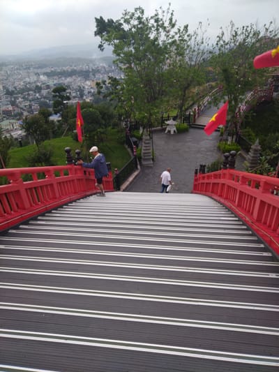 The image depicts a downward-angled view of a bridge with red railings and alternating black and white stripes on its walking surface. A man wearing a light-colored hat and a dark jacket is walking down the bridge, using the railing for support. Further down the bridge, another person in a white shirt is walking away from the viewer. At the foot of the bridge, there is a paved area with decorative elements, including a small, white, Asian-style structure. In the background, a city landscape is visible, slightly obscured by trees and vegetation. Two Vietnamese flags are placed along the bridge's path. The scene suggests a location in Hong Gai Ward, Vietnam, based on the presence of the Vietnamese flags.