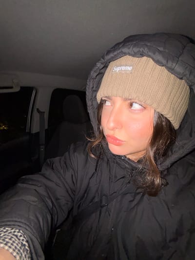 A young woman in a black hooded jacket and beige "Supreme" beanie gazes off-camera. Her lips are glossy, and she has a nose ring. The scene is dimly lit, possibly inside a vehicle, with a dark interior visible in the background. The image is a close-up, selfie-style shot angled downward.