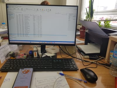 This is a medium-shot, eye-level, interior photograph of a desk with a computer monitor displaying a purchase order system. The screen shows a table with columns such as "No.", "Дата на доставка", "Vendor №", "Име", "Описание", "Кол.", "Цена", and "Статус".

In the foreground, a black keyboard is visible, with a black computer mouse to its right. A smartphone displaying a reward interface with the text "Награда 1 евро" and "Брой билети: 1" is positioned to the left of the keyboard. A blue pen lies horizontally across a piece of paper with handwritten notes. Another pen with a red cap is partially visible on the right edge of the frame.

To the right of the monitor, a Canon printer is situated, with a stack of paper in its feeder tray. Behind the printer, a clear glass vase holds several green plants, one with pink flowers, and is positioned in front of a window with white blinds.

Various office supplies, including a stapler, a small container of colorful paperclips, and a bottle of "ОстеоА" medication, are scattered on the desk. Cables are neatly bundled and run behind the monitor and printer.

The overall environment suggests an office or workspace. The image does not provide any information or visual cues that would allow for the identification of the city of Lomets, Bulgaria.
