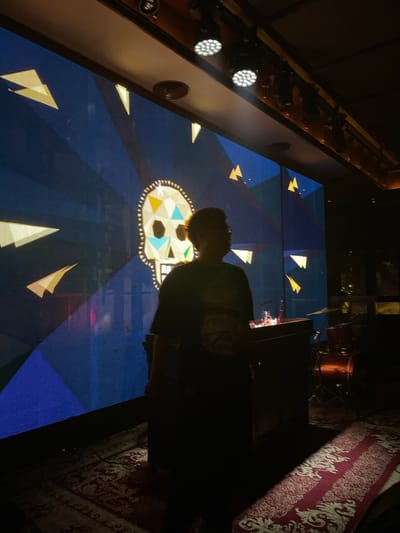 The image captures a dimly lit indoor scene, likely a performance venue or nightclub, with a large LED screen displaying a vibrant blue background adorned with yellow triangles and a central skull graphic. The skull, rendered in a colorful mosaic style, features a patterned border and geometric shapes within its eye sockets and forehead. A person, silhouetted and facing away from the camera, stands to the right of the screen, partially obscuring it. They are wearing a dark t-shirt. In the foreground, a red and white patterned carpet covers the floor. To the right, a drum set and a DJ booth or podium with some equipment are visible. The ceiling features spotlights, some of which are illuminated. The overall ambiance is dark and moody, suggesting a nighttime event. The location is Central Jakarta, Indonesia.
