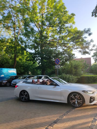 A white convertible BMW is parked on a cobblestone driveway, facing right. The car has its top down, and several people are visible inside, their heads and shoulders showing. The interior of the car appears to be a reddish-brown leather. In the background, there are several other cars parked, including a dark brown Skoda and a light blue Hyundai. A blue van with the text "Дельта-Те" is parked to the left of the other cars. A street sign with a red circle and a blue cross inside indicates no stopping or parking. Behind the cars, lush green trees form a dense canopy, with sunlight filtering through the leaves, suggesting it is daytime. The sky is a clear blue, and the overall impression is a sunny day. The setting appears to be an outdoor area, possibly a residential street or a parking lot adjacent to a wooded area. The cobblestone paving and the presence of trees suggest a well-maintained outdoor space.