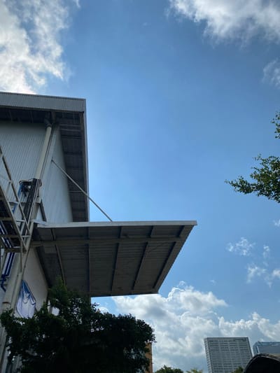 The image shows a low-angle view of a corrugated metal building with a large overhang, set against a bright blue sky with scattered white clouds. To the left, a section of a metal staircase is visible, leading up the side of the building. A person is working on the side of the building, attached to a pipe by a harness and rope. Green trees are in the foreground, partially obscuring the lower part of the building. In the distance, tall modern buildings are visible, suggesting an urban environment. The overall scene is one of industrial architecture set within a city, under a clear sky. The location is identified as Gambut Barat, Indonesia.