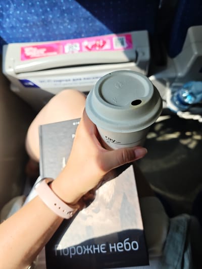 A person is holding a light gray disposable coffee cup with a black lid that has a small opening, and the cup has a logo that reads "ray cafe". The person is also holding a black book with white text that appears to be in Cyrillic. The book has the text "порожнє небо" visible on it. The person is wearing a light pink smartwatch on their left wrist. The background shows a blue patterned surface, likely a seat or upholstery, and a white and pink object, possibly part of a train or bus interior. Sunlight is casting shadows on the surface in the foreground. The scene appears to be indoors, possibly in transit. The location is not identifiable from the image itself.
