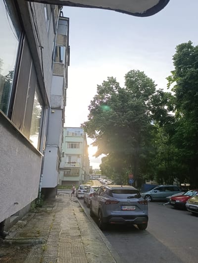 An eye-level, full shot captures a street scene in Razgrad, Bulgaria, on a bright, overcast day. The foreground is dominated by the left side of a gray, multi-story apartment building with textured walls and large windows reflecting the sky and trees. Adjacent to the building is a tiled sidewalk, which shows signs of wear and some sparse greenery.

A gray Nissan Qashqai SUV is parked on the right side of the street, facing away from the camera. Its license plate reads "PP 1233 BP". Other vehicles, including a red car and a yellow car, are parked behind it. A light blue station wagon is parked further down the street.

In the middle ground, a person is walking on the sidewalk, facing away from the camera and towards the parked cars. They are wearing a dark top and light-colored shorts. Further down the street, more parked cars are visible. A red circular "STOP" sign with an arrow is mounted on a pole to the right of the road.

The background features a row of apartment buildings, some painted in pastel colors, and a dense canopy of green trees. The sun is visible through the trees, creating lens flare and highlighting the foliage. The overall environment suggests a residential urban area.