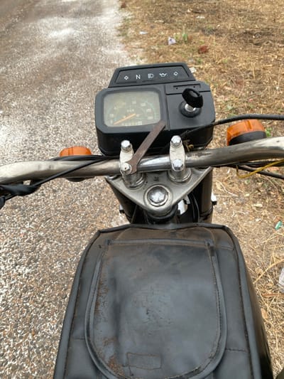 A close-up shot from above reveals the handlebars and instrument panel of a motorcycle. The panel, with its speedometer, indicators, and key, is to the left of the handlebars. A silver wrench is attached to the handlebars with metal clamps. A black, aged leather seat bag is visible to the right. The handlebars and forks are silver, contrasting with the black components. The background consists of dry grass and a gray road surface.