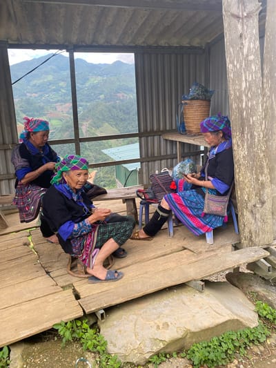 Within an open-sided, corrugated metal and wooden shelter in Dế Xu Phình, Vietnam, three elderly women are observed. The shelter features a corrugated metal roof and rear wall, wooden floorboards, and wooden support posts.

The woman on the left, positioned behind a low wooden bench, wears a dark long-sleeved top with blue accents, a plaid skirt, and a pink and blue plaid headscarf. Her hands are engaged in an activity that appears to be textile-related, possibly braiding or weaving.

The woman in the center sits on a small, round wooden stool. She wears eyeglasses, a dark long-sleeved top with blue-patterned sleeves, plaid green pants, a colorful embroidered apron-like garment, and a green and purple plaid headscarf. Her hands are occupied with a small, dark object, indicating crafting or fine motor work. Her feet are clad in blue open-toed sandals.

The woman on the right sits on a blue plastic stool, leaning against a rough-hewn wooden post. She wears a dark long-sleeved top with blue accents, a vibrant pink and blue patterned skirt, and a purple and blue plaid headscarf. A patterned shoulder bag hangs from her right shoulder. She holds and observes a silver-colored smartphone with both hands. Her left leg features a black fabric wrap or gaiter, and her foot wears a brown open-toed sandal.

On a wooden shelf behind the woman on the right, a woven basket contains a blue plastic bag filled with green, leafy material. Additional bags are visible near her stool.

The shelter overlooks a vast, verdant mountainous landscape under a partly cloudy sky. The mountains are characterized by dense foliage and visible terraced agricultural fields. A utility wire diagonally traverses the upper left portion of the background view. A structure with a green corrugated roof is visible on a distant slope, and a narrow waterfall or cascade descends down a mountain face in the lower right background.

The wooden floor of the shelter exhibits wear, with visible gaps between planks and metal support beams beneath. The ground surrounding the shelter consists of exposed soil, rocks, and patches of green vegetation.