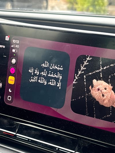 The image displays the active infotainment screen within a vehicle, observed from a slightly elevated angle. The scene is located in Qalqilya, Palestinian Territory. The screen's upper left corner shows the time "10:13" with a full "4G" mobile network signal and a high battery level icon. A vertical strip of application icons runs along the left edge, including a red-notified icon, a smiley face icon, a magenta play button, a yellow "yellow" labeled icon, a crescent moon, and a square outline.

The main display area features two rounded rectangular panels. The left panel contains white Arabic text, reading "سُبْحَانَ اللَّهِ، وَالْحَمْدُ لِلَّهِ، وَلاَ إِلَهَ إِلاَّ اللَّهُ، وَاللَّهُ أَكْبَرُ" (Glory be to Allah, and praise be to Allah, and there is no god but Allah, and Allah is the greatest). The right panel displays a light orange/cream-colored cat's face, tilted upwards and looking directly at the viewer, set against a black background with scattered white, irregular patterns. The overall screen interface background is a gradient of purple/magenta.

Below the screen, a dark, textured dashboard or center console is visible, with faint physical controls marked by horizontal white lines and symbols, suggesting volume or climate adjustments. Dust particles are present on these dark surfaces. Above the screen, through the vehicle's windshield, blurred green foliage is discernible against a lighter background, indicating an outdoor environment during daylight hours. No individuals are visible.