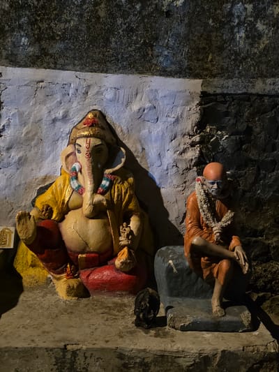 The image shows two religious statues in a dimly lit, cave-like environment. On the left is a large, seated statue of the Hindu deity Ganesha, identifiable by his elephant head and human body. The statue is adorned in yellow and red garments and has a golden crown. To the right of Ganesha, a smaller statue of Sai Baba, a revered Indian saint, sits on a dark blue stone. Sai Baba is depicted wearing an orange robe and a garland of white flowers around his neck. The background consists of a textured, crumbling stone wall. The floor appears to be made of concrete. The setting is likely a religious shrine or temple, potentially located in a more remote or natural area, given the cave-like atmosphere. While the image's context specifies Mumbai, India, this specific location within the city is not visually discernible from the image alone.