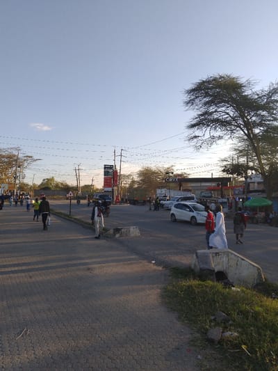 The image shows a street intersection in Athi River, Kenya, located at latitude -1.452 and longitude 36.98. The intersection is paved with asphalt and has a concrete curb on the right side. There are power lines overhead and a large tree on the right side of the image. In the distance, a gas station can be seen with a sign that reads "CIRYX". Several people are walking on the street, some wearing clothing, some wearing white robes. There are several cars parked on the side of the road, including a white hatchback. Some people are standing near the curb on the right side of the image, and some are walking in the road toward the camera. The sky is clear and blue with a few white clouds. The sun is shining, casting shadows on the ground. The environment is urban.