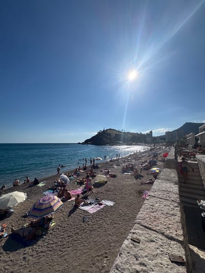 The image depicts a sunny day at a crowded beach in Sutomore, Montenegro. The beach is covered in dark pebbles, with numerous sunbathers lying on towels or seated under umbrellas. The sea is a vibrant blue, with gentle waves lapping at the shore and several boats visible in the distance. In the background, a hilly landscape with buildings and vegetation rises up, characteristic of the Sutomore coastline. The sun is high in the sky, creating bright reflections on the water and casting shadows on the beach. A stone wall with steps runs along the right side of the image, separating the beach from a walkway or restaurant area. People are engaged in typical beach activities such as sunbathing, swimming, and relaxing.