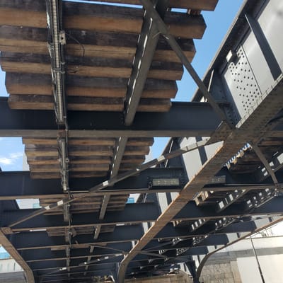 An abstract, vertically-oriented view looking upward at a structure. Dominant elements include an intricate steel framework, heavy timber shoring, and a glimpse of the sky.

The steelwork is dark and substantial, composed of vertical I-beams connected by horizontal supports. Many rivets are visible in the steel. Interspersed with the steel are rectangular timbers, likely used for shoring or support. These timbers are lighter in color, with a weathered appearance and varied wood grain.

Various cables and fixtures are suspended among the structural elements. Intermittent sections of blue sky are visible through gaps in the construction.