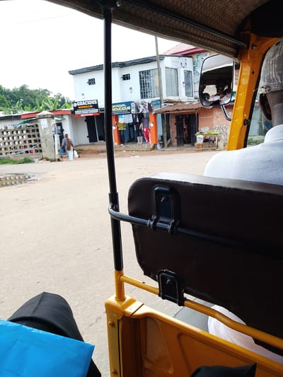The image depicts a street scene in Umudike, Nigeria, as viewed from inside a yellow auto-rickshaw. In the foreground, the dark brown leatherette upholstery of the auto-rickshaw's seat is visible, along with part of the driver's white shirt and a portion of the passenger's black trousers and a blue bag. The auto-rickshaw's yellow frame and black metal support pole are also prominent.
In the midground, a woman with long, dark hair walks away from the camera, carrying two plastic bags. She is dressed in light-colored clothing. Beside her, a white building with a red-tiled roof stands, featuring signs for "GRACIOUS PRINTING & PUBLISHING" and other businesses. A few other small shops with awnings are also visible. A metal fence with square openings is present on the left side of the scene.
The background shows more buildings, trees, and an overcast sky, suggesting it is daytime. The street itself appears to be paved with asphalt and has a few puddles. The overall environment is that of a commercial or residential area on a street in Umudike, Nigeria.