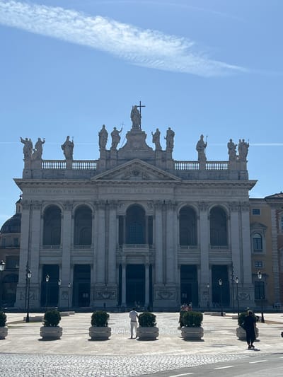 An upward-tilted, vertical photograph captures the majestic facade of a grand basilica, identified as the Archbasilica of Saint John Lateran in Rome, Italy. The building dominates the frame, showcasing its classical Baroque architecture with a prominent portico featuring large columns and arched entrances. A long balustrade crowns the roofline, adorned with numerous colossal statues of saints, with a central statue of Christ the Redeemer holding a cross, flanked by Saints John the Baptist and John the Evangelist, facing the viewer.

The sky above is a clear, bright blue with scattered wispy clouds and a faint contrail stretching across. The sunlight is strong, casting sharp shadows and illuminating the intricate details of the stone facade. In the foreground, a wide, paved plaza stretches out, with sections of cobblestones and larger flagstones. Several pedestrians are visible, walking casually across the plaza. One person in dark clothing is seen on the right, and another in white is further back. A series of large, grey planters containing green foliage are spaced along the plaza, adding a touch of greenery to the urban landscape. Street lamps are also visible on the plaza.

Visible Latin text inscribed on the frieze below the roofline reads: "CLEMENS-XII-PONT-MAX-ANNO-V" and "CHRISTO SALVATORI IN-HON-SS-IOAN-BAPT-ET-EVANG". This translates to "Clement XII, Pontifex Maximus, in the fifth year [of his pontificate], to Christ the Savior, in honor of Saints John the Baptist and John the Evangelist," confirming the building's identity and its dedication. The scene depicts a sunny day, likely midday, conveying a sense of historical grandeur and bustling city life.