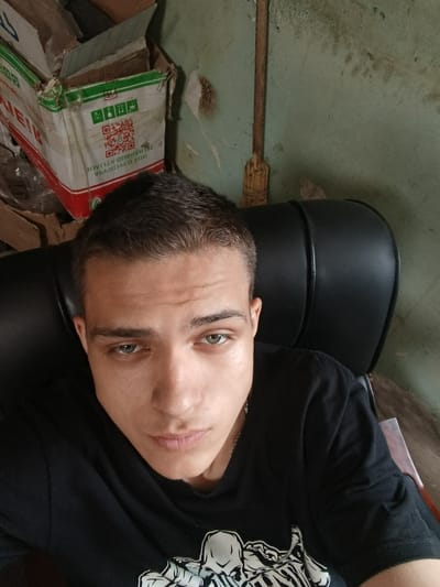A young man with short brown hair and light green eyes is shown in a close-up selfie. He is wearing a black t-shirt with a white skeleton graphic on it. He is sitting in a black chair, possibly an office chair, with a high back. The background appears to be a room with a light green, weathered wall. To the left, there is a cardboard box with red and green detailing and Cyrillic text, including QR codes. A broom with a wooden handle and straw bristles leans against the wall behind the chair. The overall impression is a somewhat cluttered indoor environment. The image was taken in Sokuluk, Kyrgyzstan.