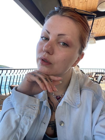A young woman is the subject of an upward-angled, medium shot taken outdoors. She has fair skin, light brown hair, and is wearing sunglasses atop her head. She wears a light blue denim jacket and a silver necklace with a red heart pendant. A tattoo is visible on her chest. Her left hand is raised, with fingers gently touching her cheek. Tattoos are visible on her hand. A black railing is visible behind her, with a view of water in the background. The location is Kunje, Montenegro.