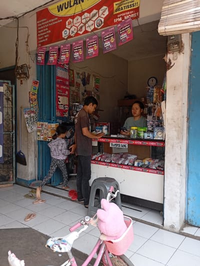 The image shows a small shop in Katapang, Indonesia (latitude: -6.99, longitude: 107.552). The shop is brightly lit and has a large sign above the entrance advertising "WULAN" and "Unlimited" mobile phone packages. The shop is filled with various products and merchandise, including phone accessories and prepaid cards.
Two people are present in the shop: a man in a blue shirt and jeans, standing near the counter and looking at something in a shelf, and a woman in a blue shirt and black skirt, standing behind the counter and looking at something on her phone.
A small child, wearing a pink shirt and blue shorts, is standing in the left side of the image, looking at something in a display rack behind the counter. The child is wearing a backpack and holding a red bag in their right hand.
The image is taken from the left side of the shop. The floor is tiled and clean. A small pink and white bicycle is partially visible in the bottom right corner.