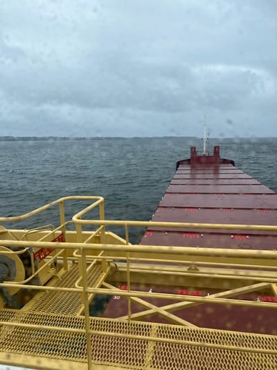 The image presents a forward-facing view from a large vessel off the coast of Ploemeur, France, under an overcast sky. In the immediate foreground, to the left, a yellow-painted metal structure is visible, featuring grated platforms and guardrails. This structure incorporates a partially visible cylindrical mechanism, possibly a winch, and a red and white label marked "SWL". The vessel's dark red main deck extends from the midground towards the horizon, composed of multiple large, flat rectangular panels, likely cargo hatch covers. A white mast-like structure is visible at the furthest end of the visible deck. The vessel is navigating dark, choppy water. In the distant background, a low, indistinct coastline with sparse structures or vegetation is visible. The sky is uniformly grey and cloudy. Numerous water droplets are present on the transparent surface through which the photograph was taken, indicating rain or sea spray. No individuals are depicted, and no specific actions or interactions are occurring.