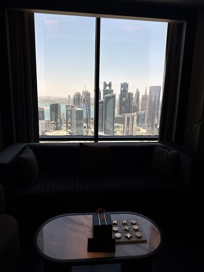A window in a high-rise building in Doha, Qatar, overlooks the city skyline. The view includes various skyscrapers, water features, and urban landscaping. The window is divided into panes by dark vertical and horizontal lines. Heavy dark drapes frame the window. To the right of the window, a dimly lit interior features a ridged sofa with pillows and a side table holding a tic-tac-toe board game. The lighting is dim in the interior, contrasting with the bright outdoor scene visible through the window.