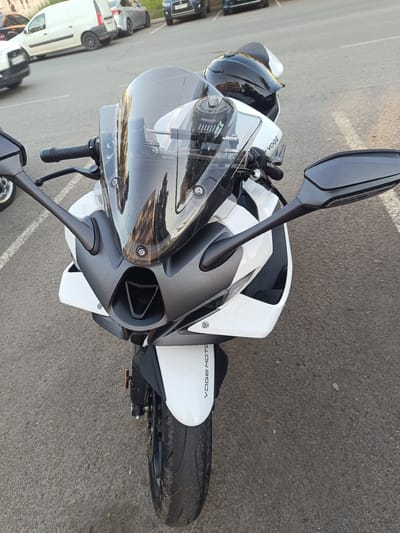 The image shows the front of a white and grey sport motorcycle parked on an asphalt surface, possibly a street or parking lot, in Naberezhnye Chelny, Russia. The motorcycle has a dark tinted windshield, black handlebars, and mirrors. A black helmet is visible on the seat of the motorcycle. In the background, several parked cars and a white van are visible, along with markings on the asphalt that suggest a parking area. The lighting suggests it is daytime, likely late afternoon or early evening due to the warm tones in the reflection on the windshield. There are no people visible in the image. The overall scene depicts a parked motorcycle in an urban environment.
