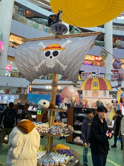 This media file captures an indoor commercial space, likely a shopping mall or a themed exhibition area in Guang Zhou Shi, China, dedicated to the popular anime "One Piece." The image is presented in a vertical, portrait orientation.

In the mid-ground, a prominent, large-scale replica of a pirate ship's sail is featured, displaying the iconic Straw Hat Pirates Jolly Roger (a skull and crossbones wearing a straw hat). This sail is attached to a wooden mast structure. To its left, a smaller black flag also bears a pirate skull design. Above the sail and to the left, a large, bright yellow, circular inflatable object is visible, possibly part of a hot air balloon or another decorative element.

Several people are present, indicating a public event or shopping activity. In the mid-ground, two young men with dark hair are looking towards the camera. The man on the right wears a black hoodie, and the one behind him wears a dark blue hoodie. In the foreground, a person in a cream or white puffer jacket is seen from behind, looking at merchandise. Other blurry figures can be discerned in the background.

The area is filled with One Piece-themed decorations and merchandise. Behind the people, cardboard standees of various One Piece characters, including Monkey D. Luffy, Roronoa Zoro, and Sanji, are set up. A multi-level structure with a distinctive red and yellow striped dome top resembles a building from the series. To the right, multiple merchandise stalls are laden with numerous plush toys of various colors like purple, yellow, white, and grey. A large white plush head, possibly a character, is also visible.

The activity appears to be people browsing and enjoying a themed retail or promotional event. The setting suggests a vibrant atmosphere with bright indoor lighting. Visible text includes "Rulun" and "Totor" on small flags, and "SANJI" on a character cutout. The overall scene highlights a lively fandom event.