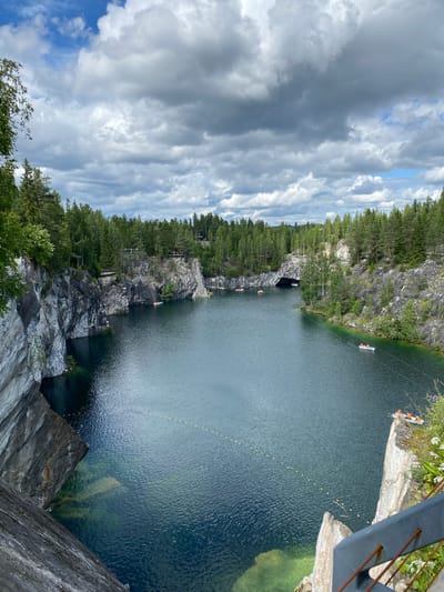 The image depicts the Ruskeala Mountain Park, a former marble quarry in Karelia, Russia, featuring a serene lake surrounded by towering, steep marble cliffs. The water is a deep, clear blue-green, reflecting the dramatic cloudy sky. Several small boats are scattered across the lake, some with occupants wearing orange life vests, suggesting recreational activity. Lush green trees and dense coniferous forests cover the tops of the cliffs and extend into the background, creating a stark contrast with the rocky terrain. In the distance, a cave-like opening is visible in the cliff face. The foreground shows a section of a grey marble cliff face with visible strata, and a metal railing suggesting a viewing platform or walkway. The overall impression is one of natural beauty and tranquility, with a hint of adventure.