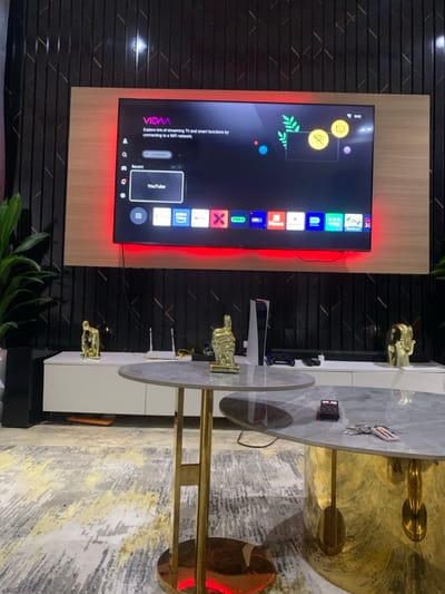 A modern entertainment setup features a large flat-screen TV mounted on a light wood panel, accented with red LED strip lighting. The TV displays a user interface with various app icons, including YouTube. A contemporary coffee table with a light gray marble top and brass legs is adorned with golden figurines. The surrounding decor includes black textured wall panels and a gray patterned rug, suggesting a stylish and well-designed interior space.