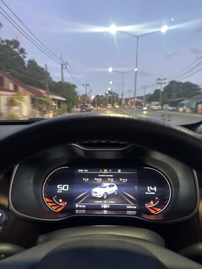 This media file depicts the interior of a car, looking out through the windshield and at the car's digital dashboard. The dashboard displays various driving information including speed, fuel efficiency, distance traveled, and trip details. A prominent element is the digital speedometer showing "50 km/h". Other visible text on the dashboard includes "Current Journey", "1.8 km", "00:10 h:min", "B152 km", "10 km/h", "11.3 km/L", "Long Press OK to Reset", and "53434 km". It also shows "1.4 UminOX000".
The scene outside the car appears to be a road in Kathu, Thailand, at dusk. The sky is a mix of blue and gray, with bright lights from street lamps and streetlights suggesting twilight. There are power lines and trees visible, typical of a semi-urban or suburban setting. The road seems to be a two-lane street with some traffic, including other vehicles and possibly motorcycles. The overall impression is that of driving during the transition from day to night, with ambient light casting a soft glow. There are no people clearly visible in the shot. The focus is on the car's interior and the driving experience it represents.