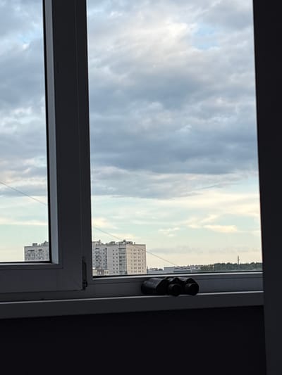 The image shows a view through a window of a cloudy sky and some buildings. Binoculars are visible on the windowsill. The sky is filled with gray and white clouds, with some patches of lighter sky visible. In the distance, there are several tall buildings, some of which appear to be apartment complexes. A line of trees is also visible in the background. The overall impression is of an overcast day, likely in an urban or suburban setting. The time of day appears to be either early morning or late afternoon, given the soft lighting. The image is taken from a high vantage point, suggesting the view is from an upper floor of a building. There is some faint text on a building in the distance, but it is too blurry to be legible. The scene is static and there is no discernible activity taking place. The mood is calm and somewhat somber due to the cloudy sky.