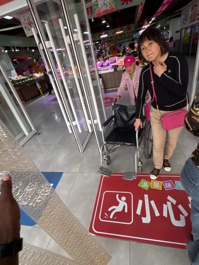 A medium-angle shot reveals a woman in a black jacket and beige pants sitting in a wheelchair, accompanied by another woman wearing a pink hat. The wheelchair is positioned on a gray-tiled floor, and a red sign with white Chinese characters and a running person symbol is visible on the floor near the wheelchair. The wheelchair user is looking towards the camera. Transparent barriers extend from the bottom left towards the center of the frame, showing a shop interior with various products. A hand holding a white decorative panel is visible in the lower right corner. The image was likely taken in a commercial setting in the city of Guta, China.