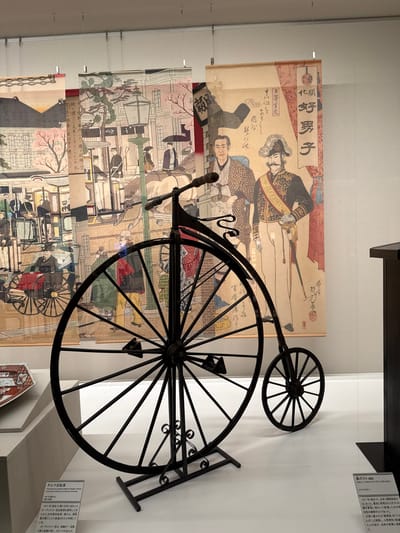 The image captures a museum display featuring historical artifacts. Prominently displayed is a replica of a penny-farthing bicycle, characterized by its large front wheel and smaller rear wheel. Behind the bicycle hang several antique Japanese prints depicting scenes of daily life, including portraits of men in traditional attire, streetscapes with horse-drawn carts, and individuals interacting in public spaces. Text panels with Japanese characters are positioned near the bicycle and to the right, providing explanations and context for the exhibits. The setting is the Sumida River Museum in Sumida City, Japan.