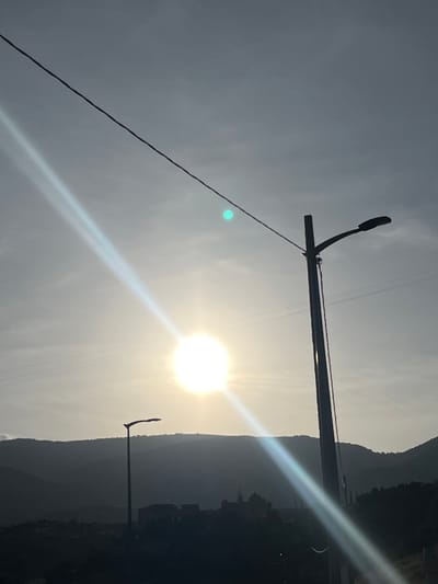 The image depicts a landscape in Ouaouizert, Morocco, possibly during sunrise or sunset, given the low angle of the sun. The sun is obscured by the light, however, there are a set of hills to the right. Two power poles are visible, one in the foreground and one in the background. A power line stretches across the top left of the image. The sky is overcast. The image is shot at an angle.
