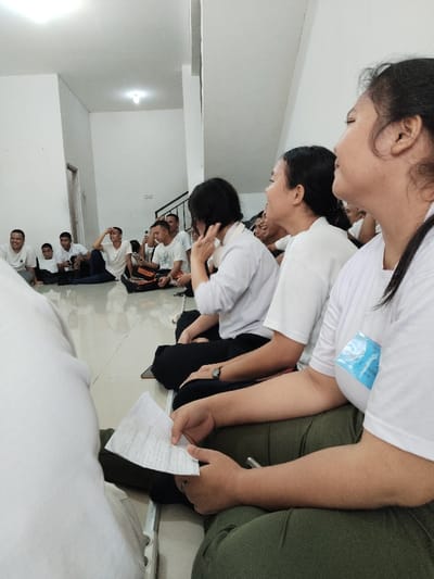 The image shows a group of young adults sitting on the floor of a room in Telukjambe, Indonesia, located at the coordinates -6.308 latitude and 107.282 longitude. The room has white walls and a white ceiling. The floor is tiled. Several of the individuals are wearing white shirts, and one individual is holding a piece of paper in their hands. The scene appears to be a casual gathering of friends. One of the individuals, a young woman with long black hair, is seated with her right arm outstretched, holding a piece of paper in her hand. Her gaze is directed towards the group of individuals. The other young adults are seated in a circle, and they appear to be talking to each other.  There is a light source in the ceiling, and a set of stairs is visible in the background. The image is taken from a low angle, and the focus is on the young woman holding the paper. The scene suggests a friendly and informal interaction.