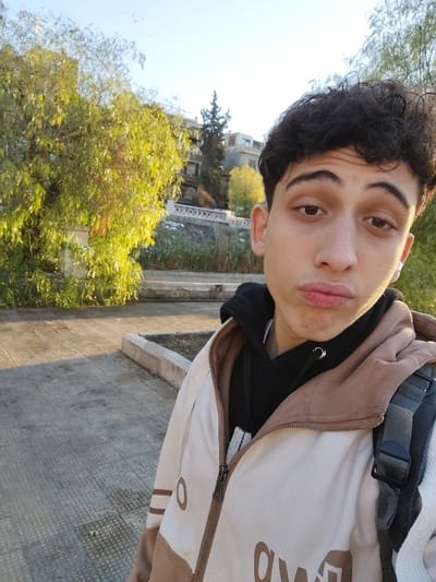 The image captures a close-up outdoor portrait of a young male, appearing to be in his late teens or early twenties, within As-Salihiya Municipality, Syria. He has dark, curly hair and light brown eyes, looking directly at the camera with a slight pout. He is dressed in a black hooded sweatshirt layered under an unzipped, two-tone jacket that is cream/off-white on the upper portion and light brown on the lower. Partial dark lettering is visible on the jacket. A dark-colored backpack with a gray strap is visible on his right shoulder, and he wears a small silver hoop earring in his left ear and a white earbud in his right ear.
The background features a paved ground surface with a low concrete barrier on the right side. Behind the subject, a line of trees with green and yellowish foliage is visible, illuminated by what appears to be morning or late afternoon sunlight, casting long shadows. Further back, a stone retaining wall topped with a white decorative balustrade is present, with more vegetation below it. Beyond the wall, multi-story buildings and a tall, dark coniferous tree are visible on an elevated terrain against a clear, pale blue sky.