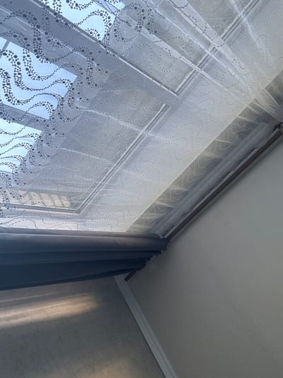 The image shows a close-up view of a window covered with sheer white curtains adorned with a wavy pattern of small dark dots. The curtains are translucent, allowing light to filter through from the outside, revealing the window panes. To the right of the sheer curtains, a darker, solid curtain is partially visible, suggesting a layered window treatment. The scene is set indoors, with a view of the ceiling and a wall in the background. The ceiling is white with molding, and the wall has a textured finish. A strong beam of sunlight is casting a distinct shadow or reflection on the wall, indicating that it is daytime, likely late afternoon or early morning when the sun is at a low angle. There is no visible text in the image. The overall impression is one of a quiet, domestic interior, with natural light playing a significant role in the composition. The location context of Яромінскі сельскі Савет, Belarus, suggests a rural or village setting.