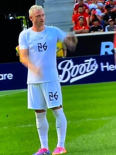The image depicts a male soccer player on a green grass field within an outdoor sports stadium, specifically located in the city of Casablanca, Morocco. The player, with short blonde hair and fair skin, is positioned centrally, wearing a white short-sleeved athletic shirt and matching white shorts, both prominently displaying the stylized black number "26". He wears white athletic socks with a black Nike swoosh visible on his left leg, and multi-colored (pink and blue/purple) soccer cleats. His right arm is partially blurred, suggesting motion. In the background, seated spectators are visible in stadium stands, predominantly wearing red attire. Below the spectators, a dark blue advertising board is visible, featuring white text including "are", the prominent "Boots" logo in cursive script, and partially visible "He" (likely part of "Health") and a large "R". The scene is brightly lit.